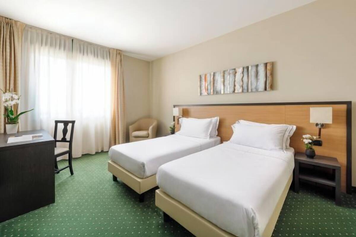A hotel room with two beds, a desk, a chair, a nightstand, and a painting on the wall.