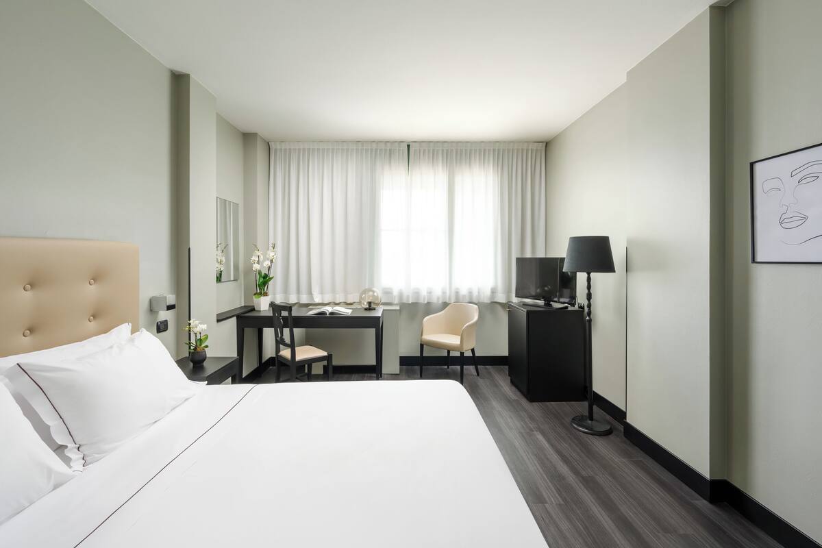 A hotel room with a large bed, a desk with a chair, a television, and a window with curtains.