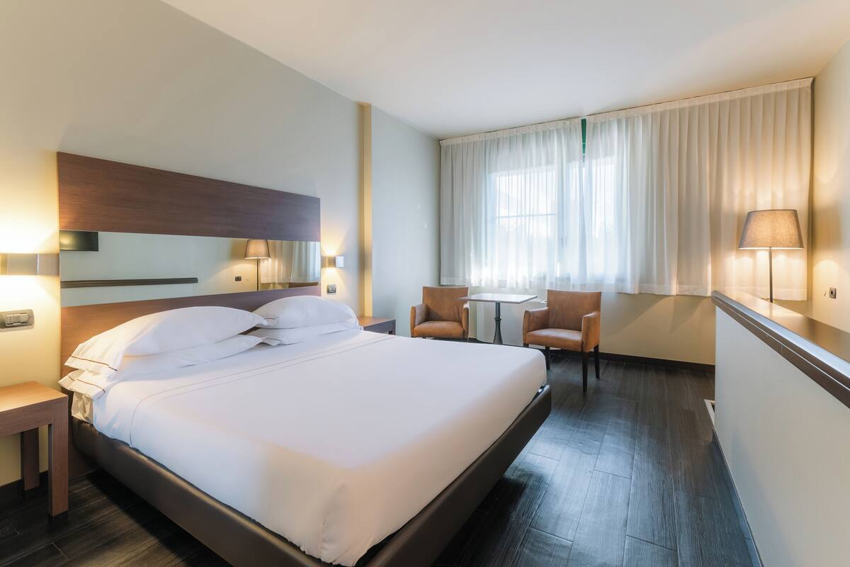 A hotel room with a large bed, two bedside tables with lamps, a small table with chairs, and a window with curtains.