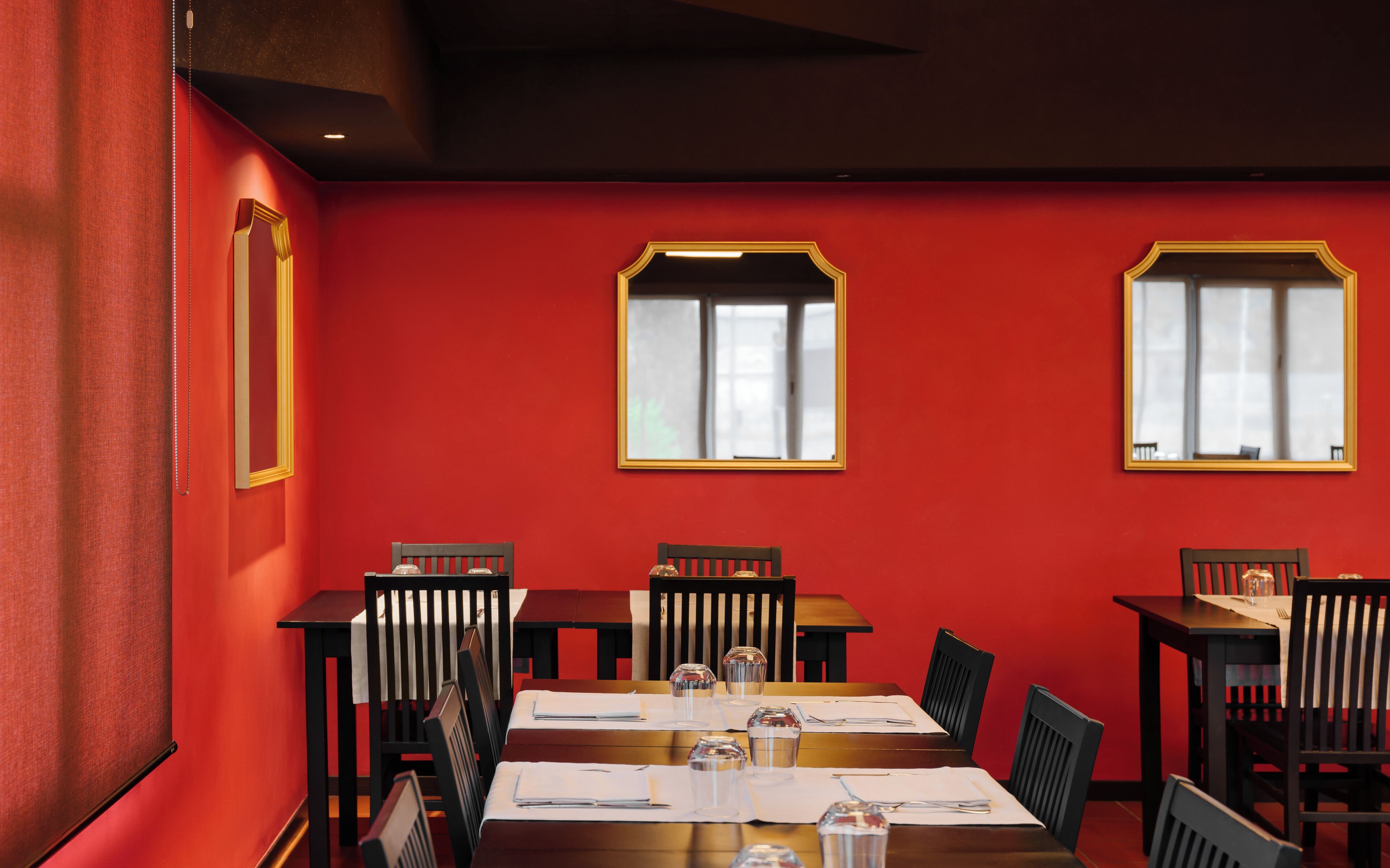 A dining area with red walls, wooden tables, and chairs. There are two windows with white curtains.