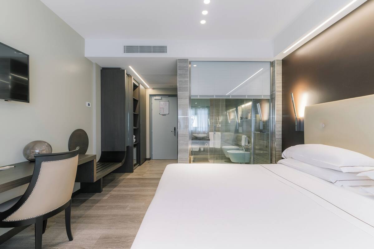 A modern hotel room with a large bed, a desk with a chair, a TV, and a glass-enclosed bathroom.