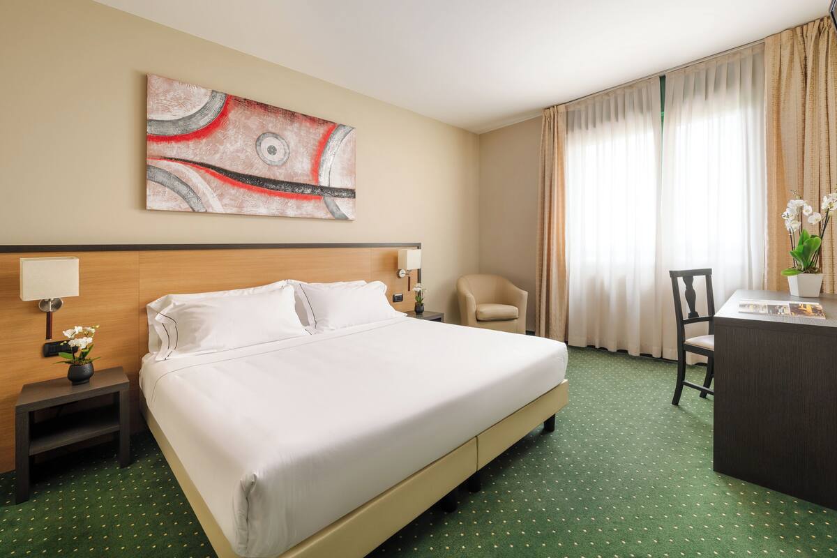 A hotel room with a large bed, a desk, a chair, a nightstand, and a painting on the wall.