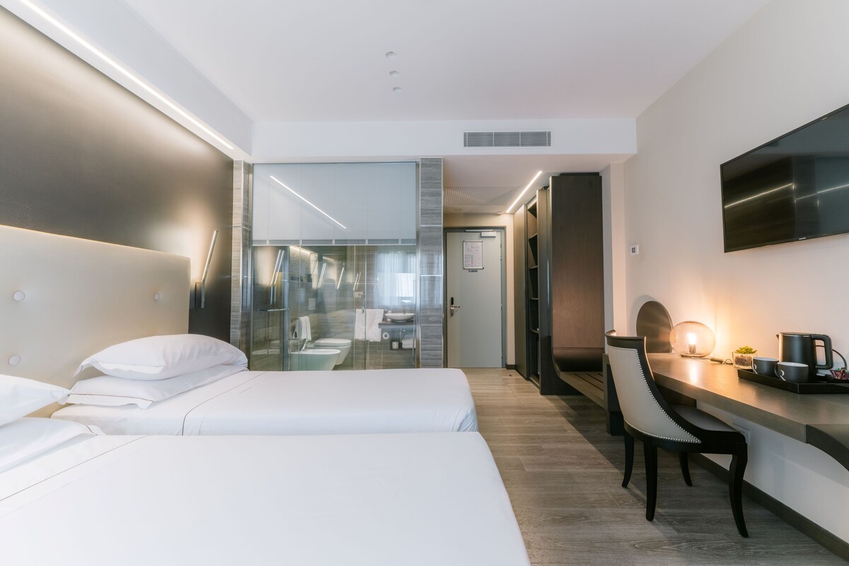 Une chambre d’hôtel avec deux lits, un bureau, une chaise, une télévision et une salle de bain visible à travers une paroi en verre.