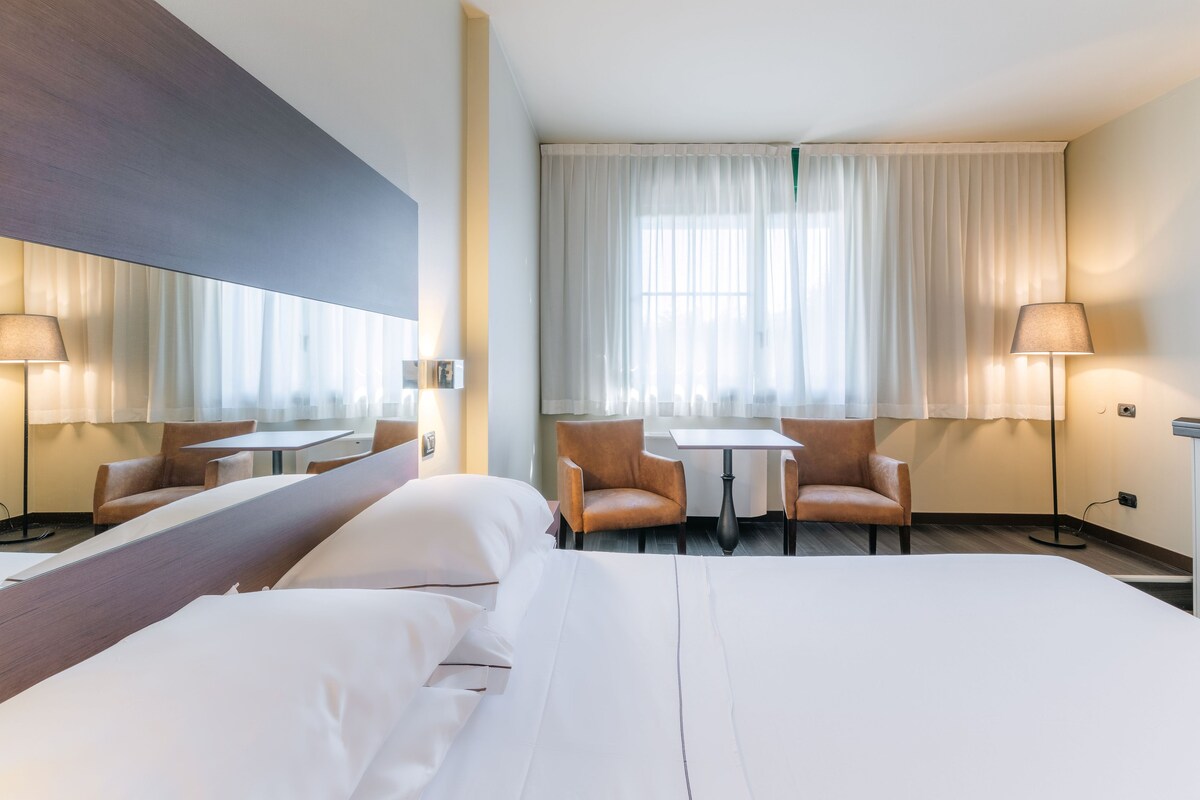 Une chambre d’hôtel avec un grand lit, deux fauteuils, une petite table et une grande fenêtre avec des rideaux.