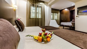 A hotel room with a bed, a bedside table with a lamp, a fruit arrangement on the bed, a desk with a chair, and a TV mounted on the wall.