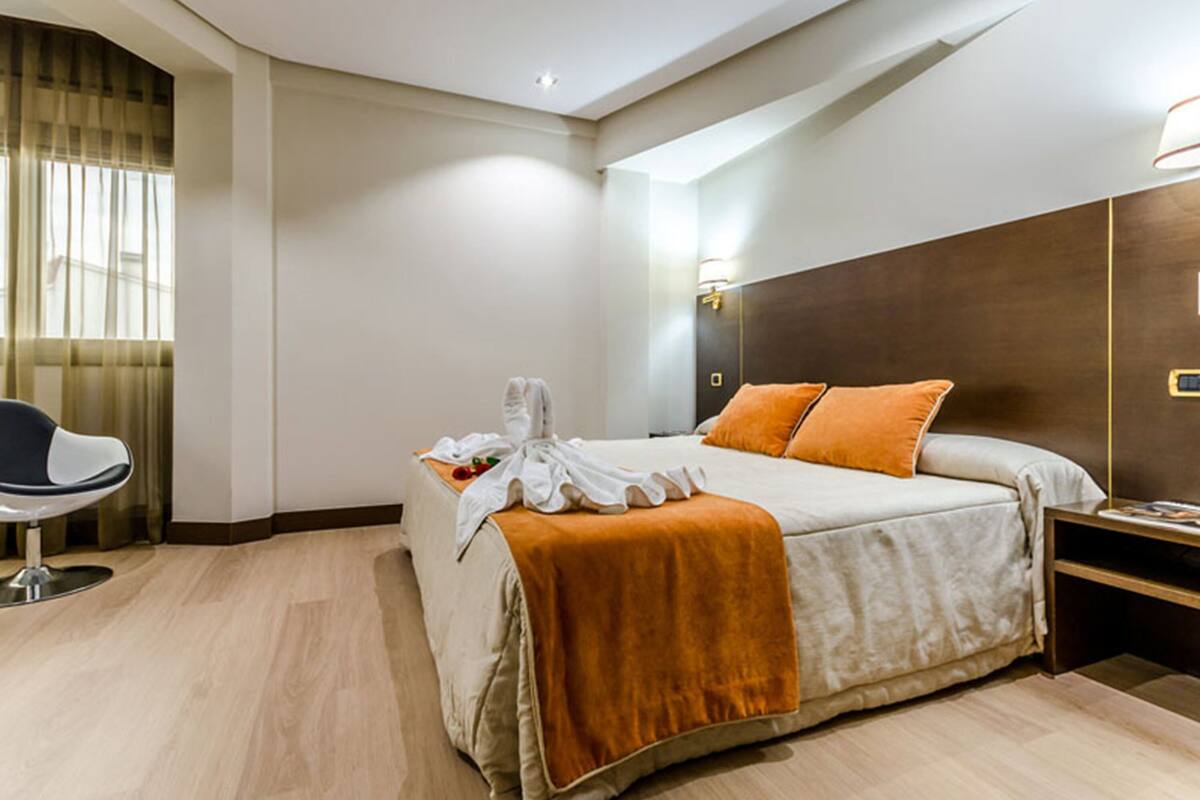 A hotel room with a bed, a chair, a nightstand, and a wall-mounted lamp.