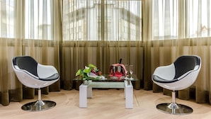 A room with two modern chairs, a glass table with a red bag and a bottle, and a view through sheer curtains.