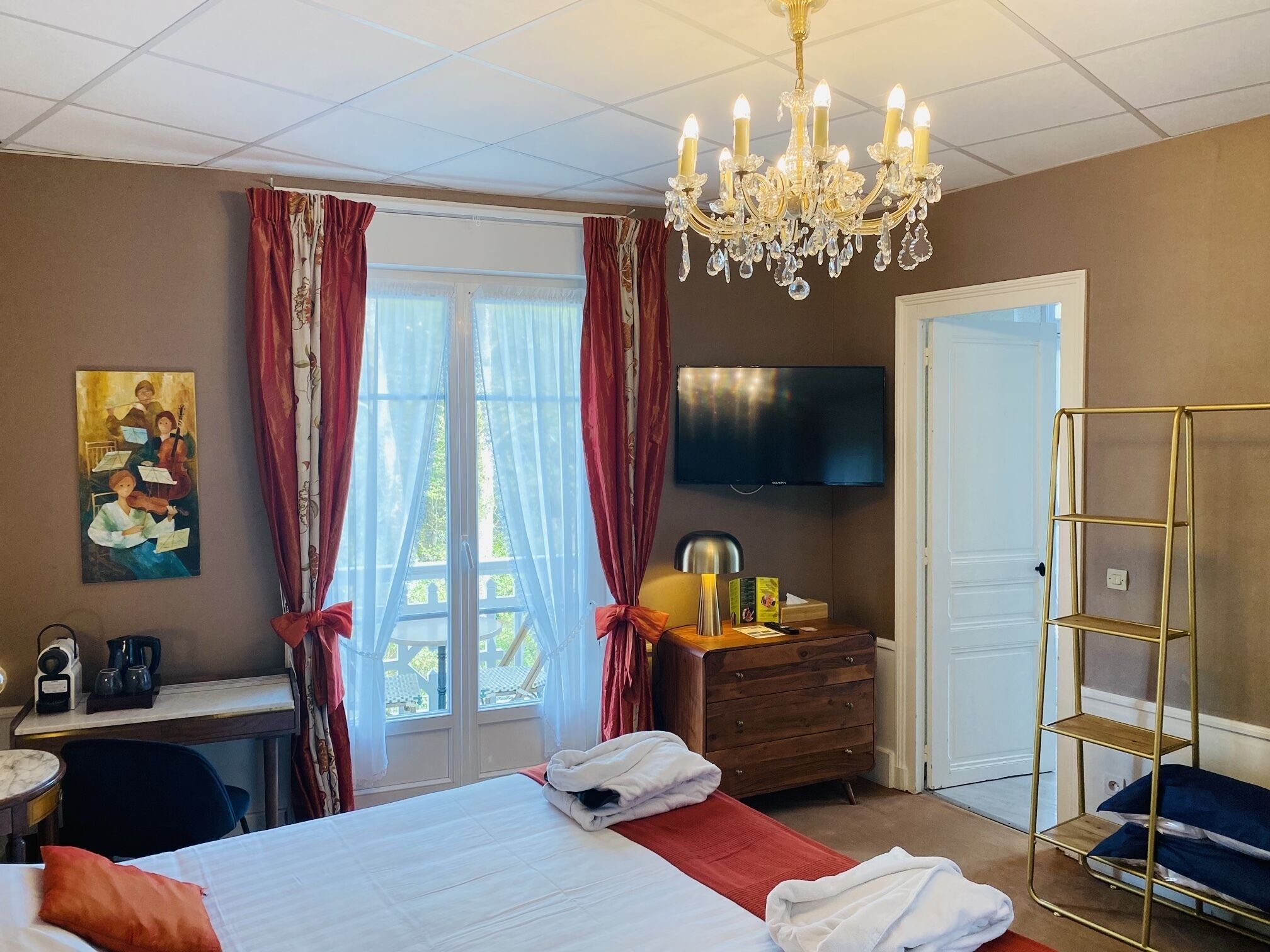 A hotel room with a large bed, a desk with a chair, a TV mounted on the wall, and a chandelier.