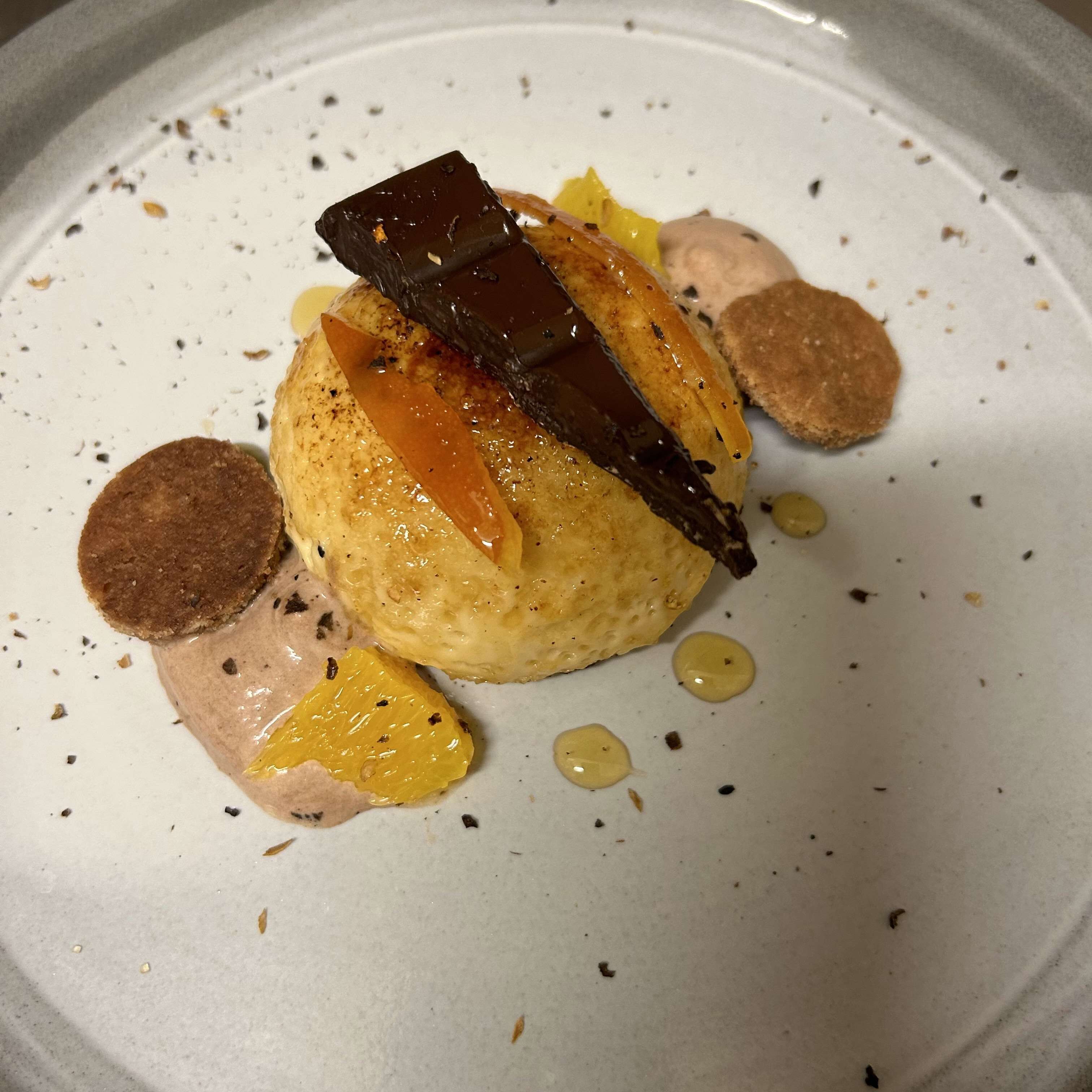 A dessert with a round base, topped with a chocolate piece and orange slices, accompanied by small round items and a liquid component.