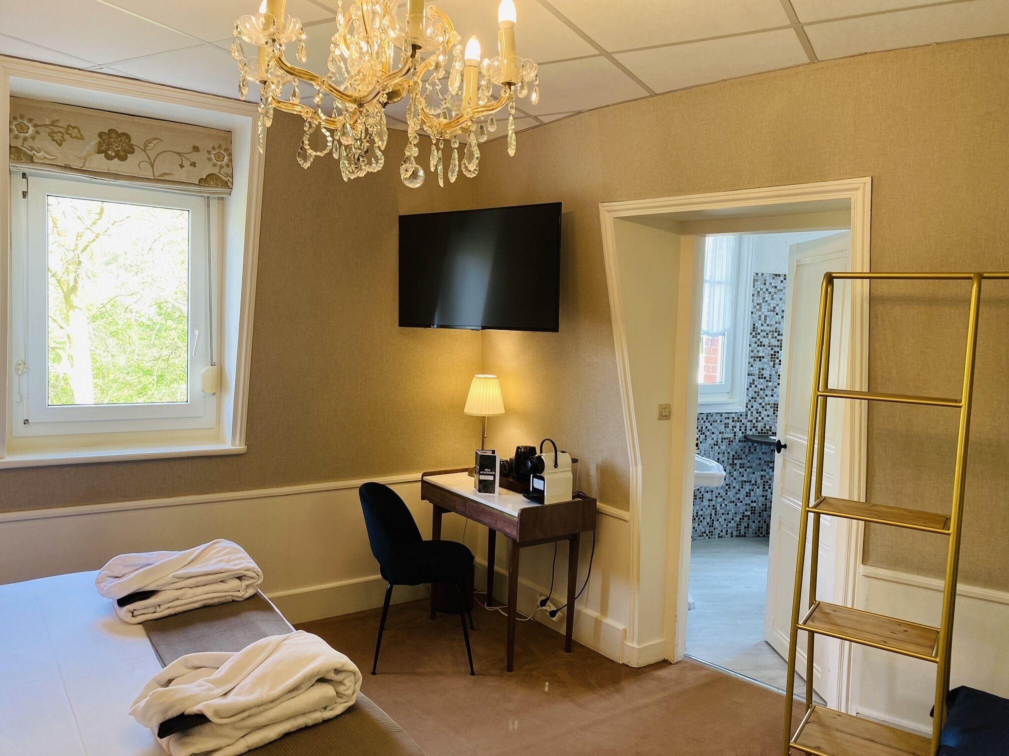 Habitación de hotel con cama, escritorio con silla, televisor, candelabro y vista al exterior.