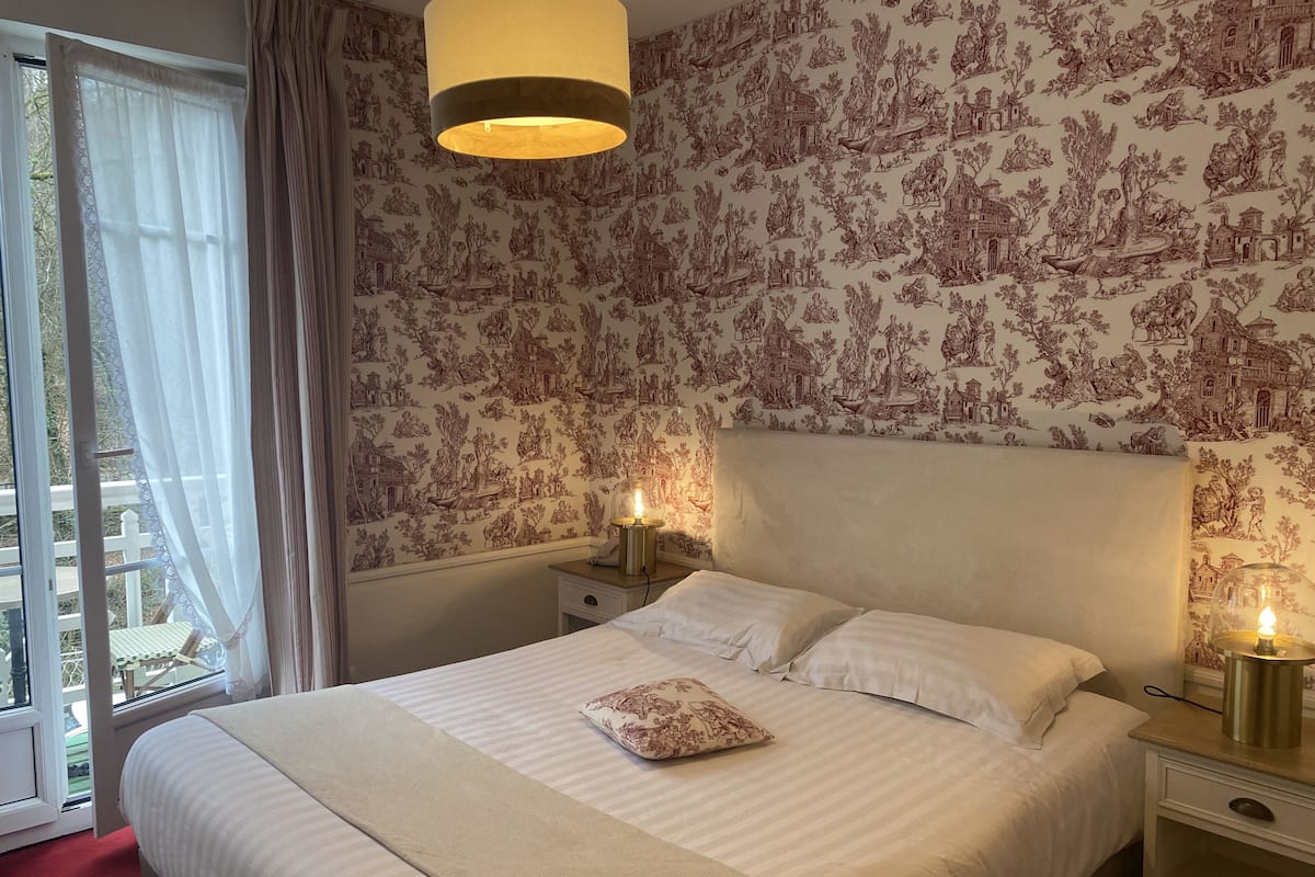 Une chambre d’hôtel avec un grand lit, deux tables de chevet avec des lampes, une fenêtre avec des rideaux et un papier peint à motifs.