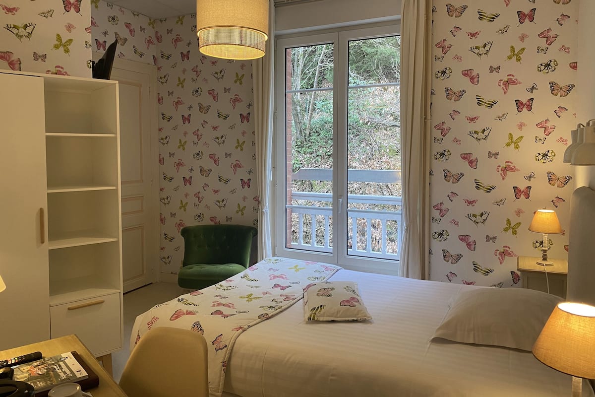 Une chambre à coucher avec un lit, un fauteuil vert, une armoire, une fenêtre donnant sur des arbres et un papier peint orné de motifs de papillons.