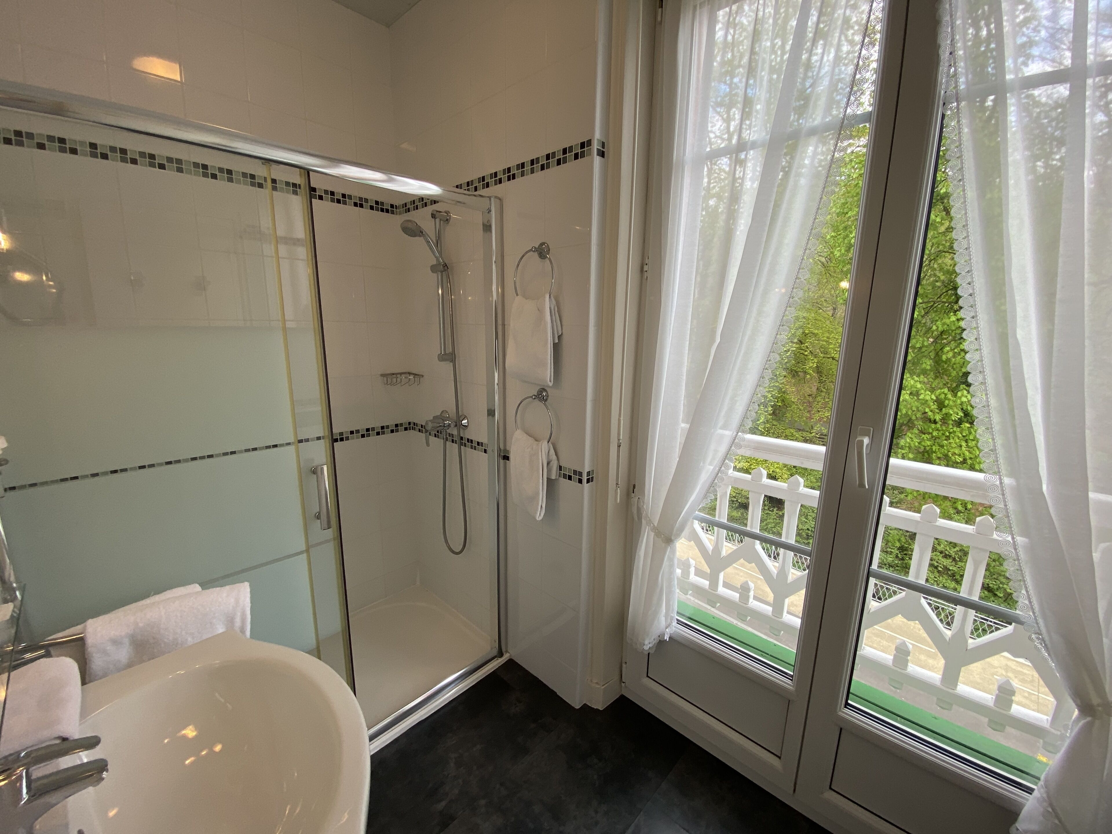 Une salle de bain moderne avec une douche en verre, un lavabo blanc et une vue sur un balcon agrémenté d’une balustrade blanche.