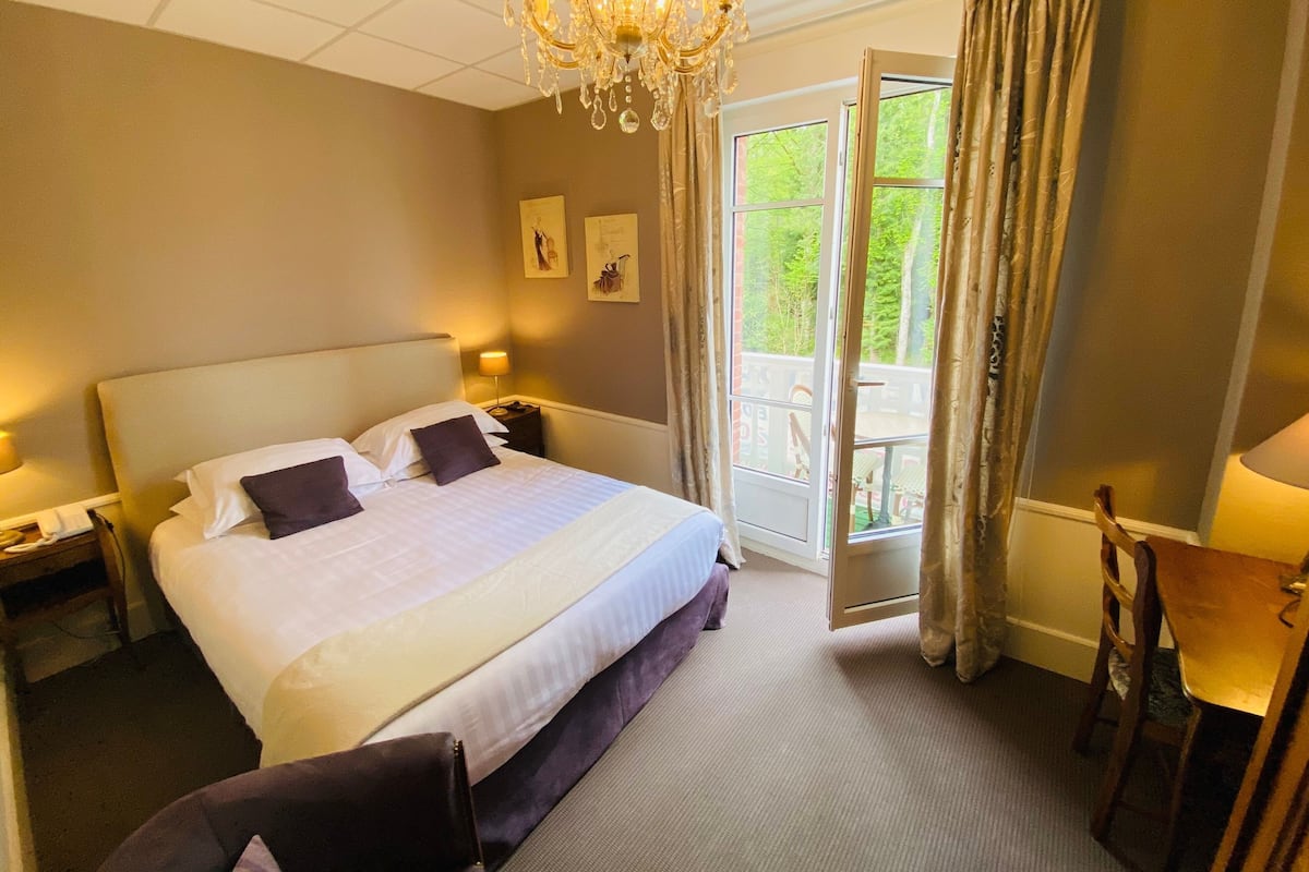 Une chambre d’hôtel avec un grand lit, des lampes de chevet, un lustre, un balcon avec des rideaux et une vue sur la verdure.