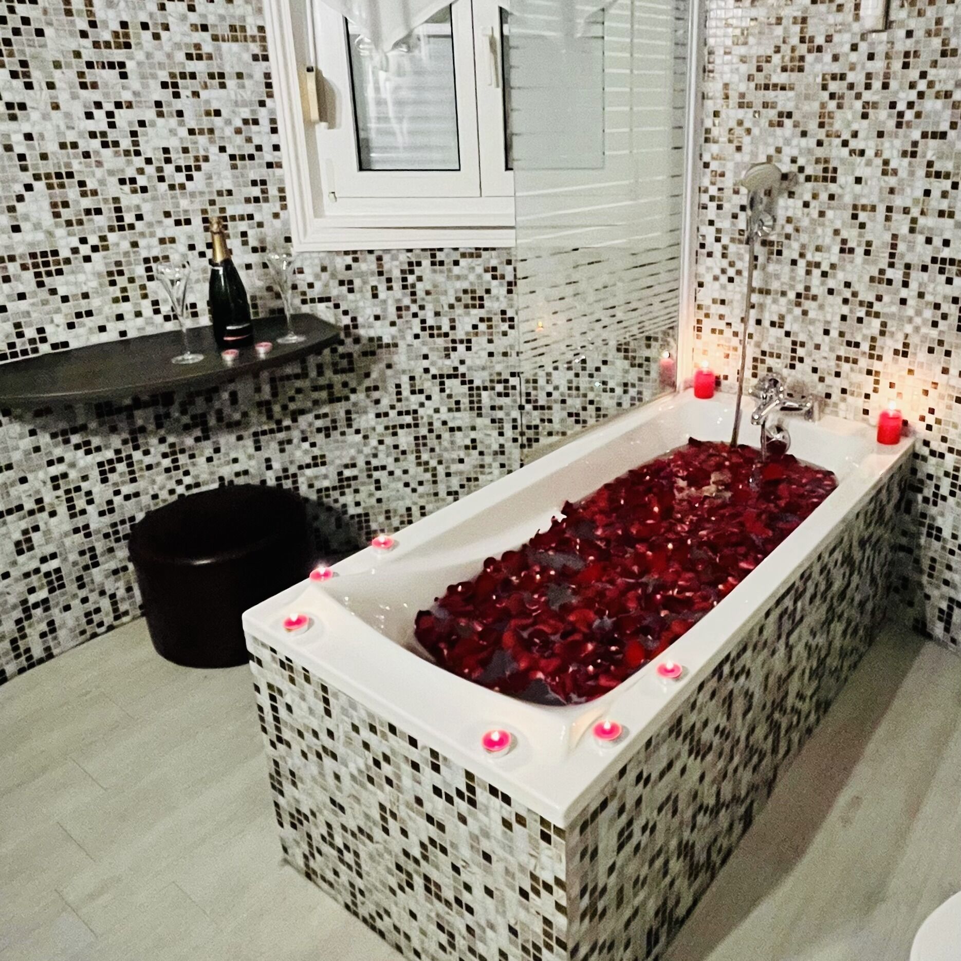 A bathroom with a mosaic-tiled wall, a bathtub filled with rose petals, a bottle of champagne, and candles.