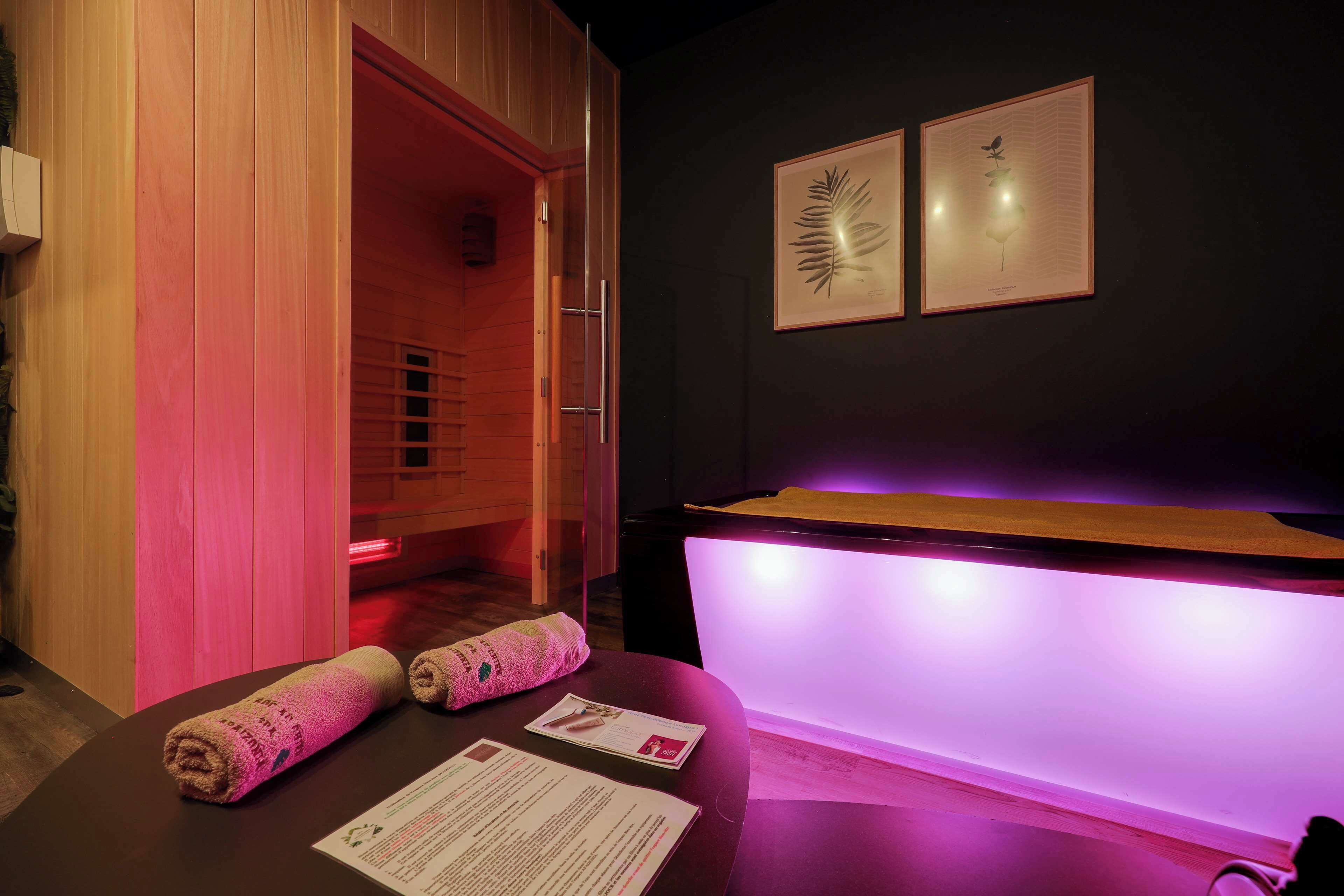 Um quarto de spa com porta de madeira, um balcão de recepção com iluminação roxa e duas gravuras botânicas emolduradas na parede.