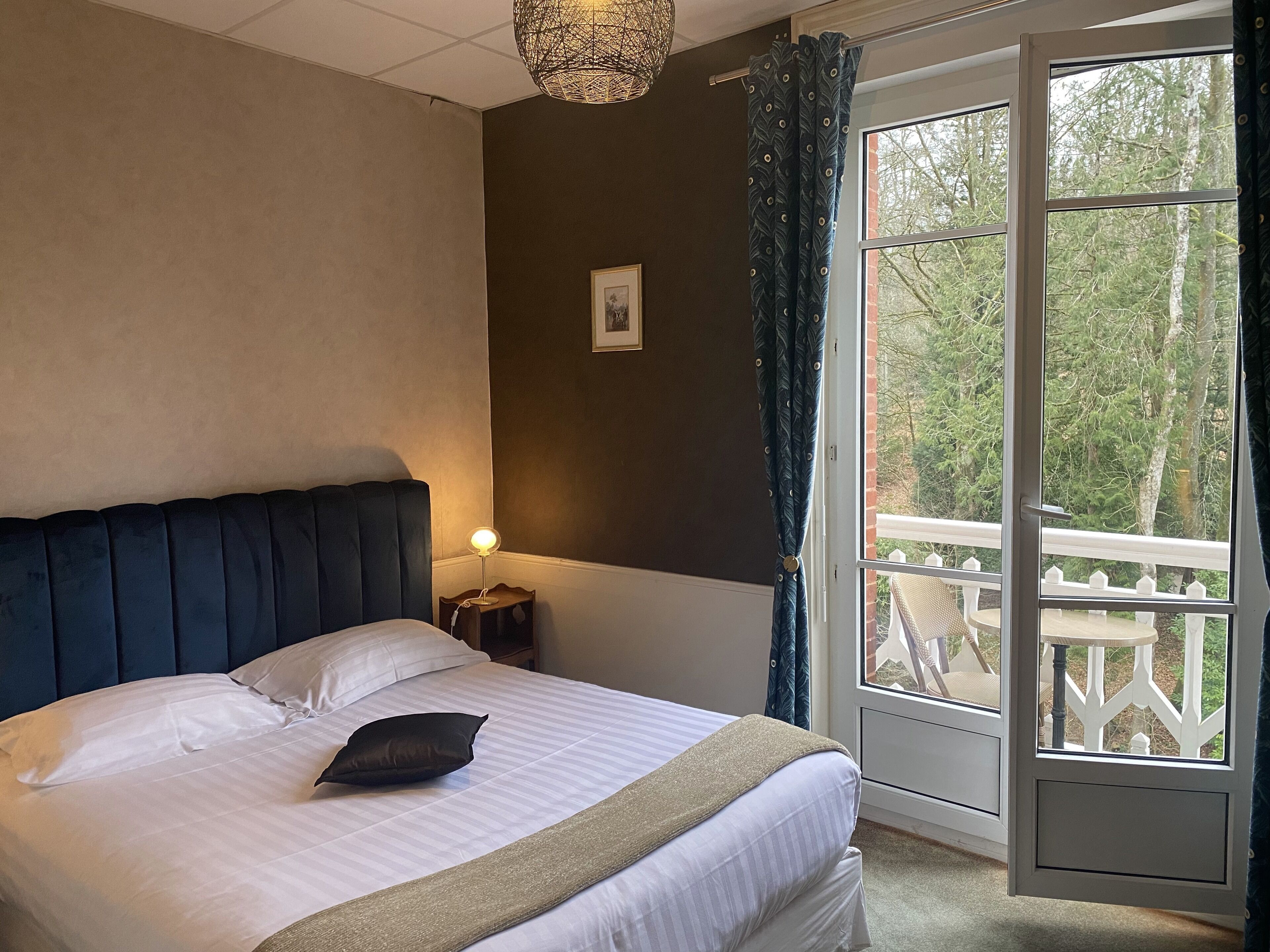 A bedroom with a bed, a bedside lamp, a picture on the wall, and a view of trees through a window.