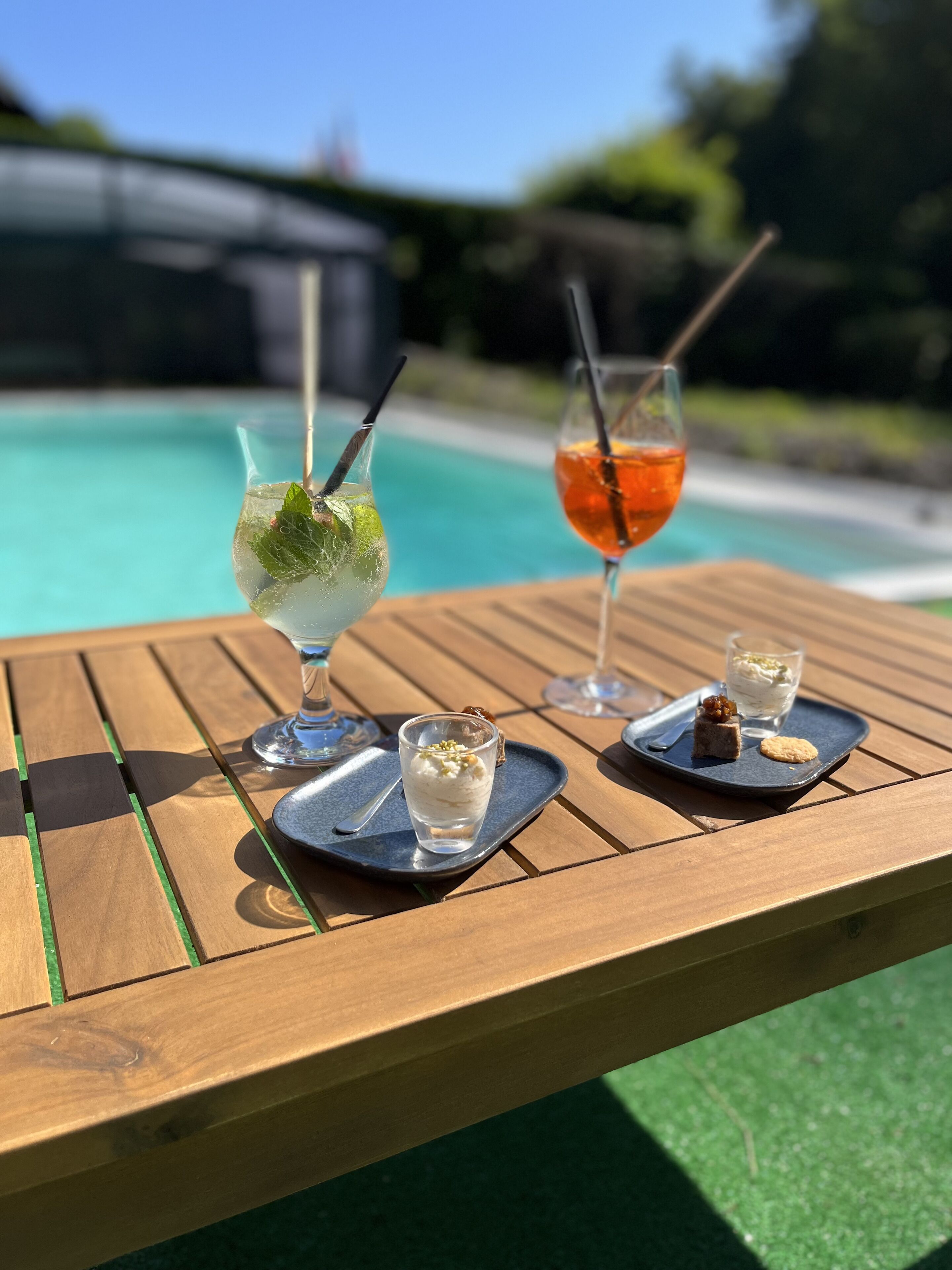 A wooden table with a glass of iced drink garnished with mint, a glass of orange cocktail, and a dessert tray with a scoop of ice cream and a cookie.