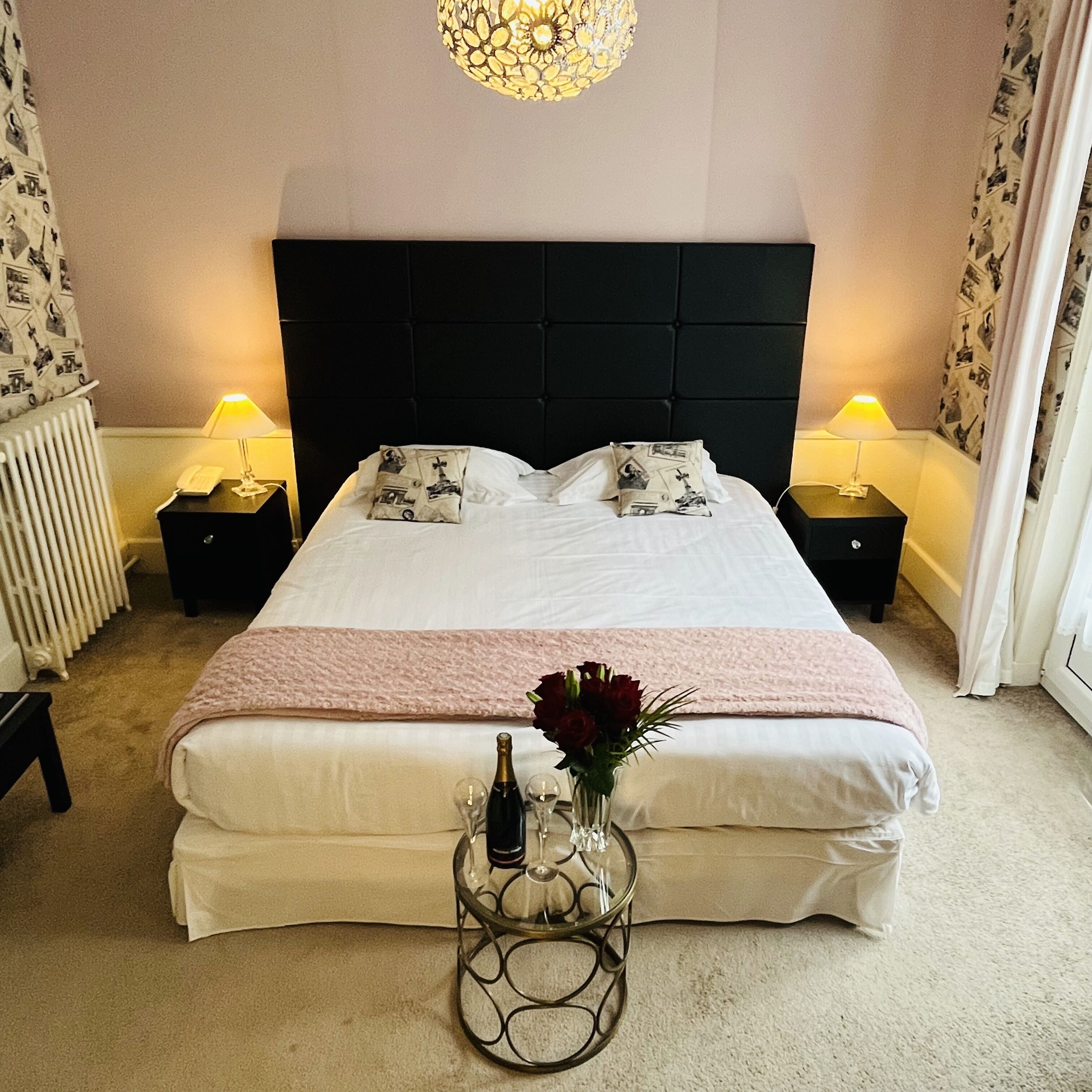 A neatly made bed with a pink blanket, two bedside tables with lamps, a glass table with a bottle and glasses, and a wall with patterned wallpaper.
