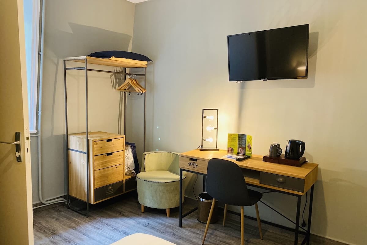 Une pièce de taille modeste, comprenant un bureau en bois, une chaise, une petite table ronde, un luminaire suspendu et un téléviseur à écran plat fixé au mur.