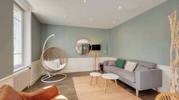 Una sala de estar con un sofá gris, un sillón de cuero marrón, un sillón de mimbre, un espejo redondo y una mesita.