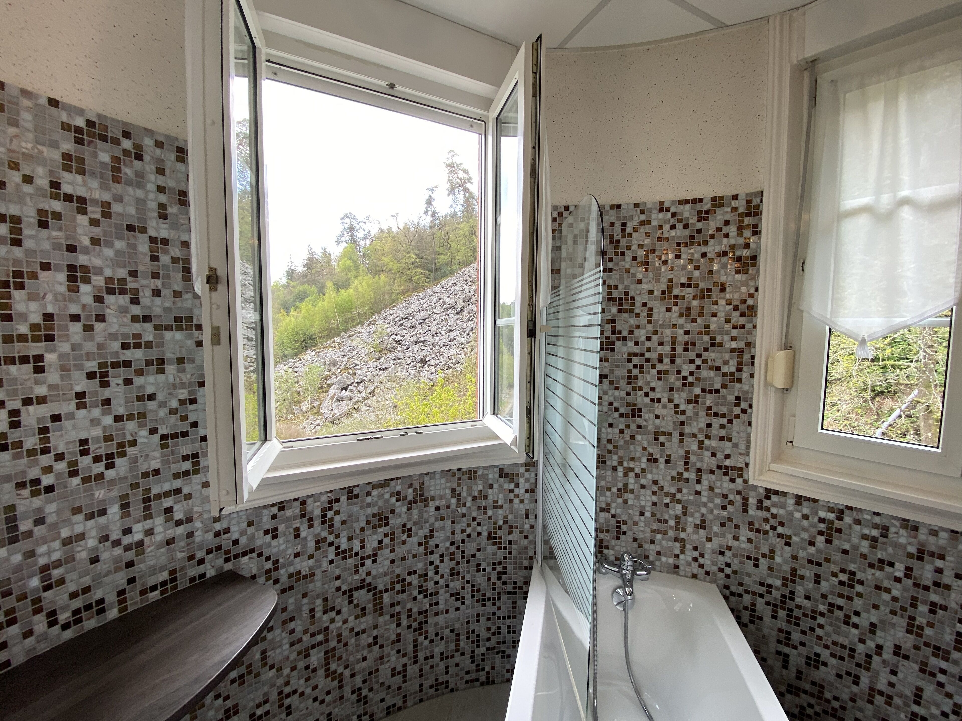 A bathroom with mosaic tile walls, a bathtub, and a window with a view of a rocky hillside.