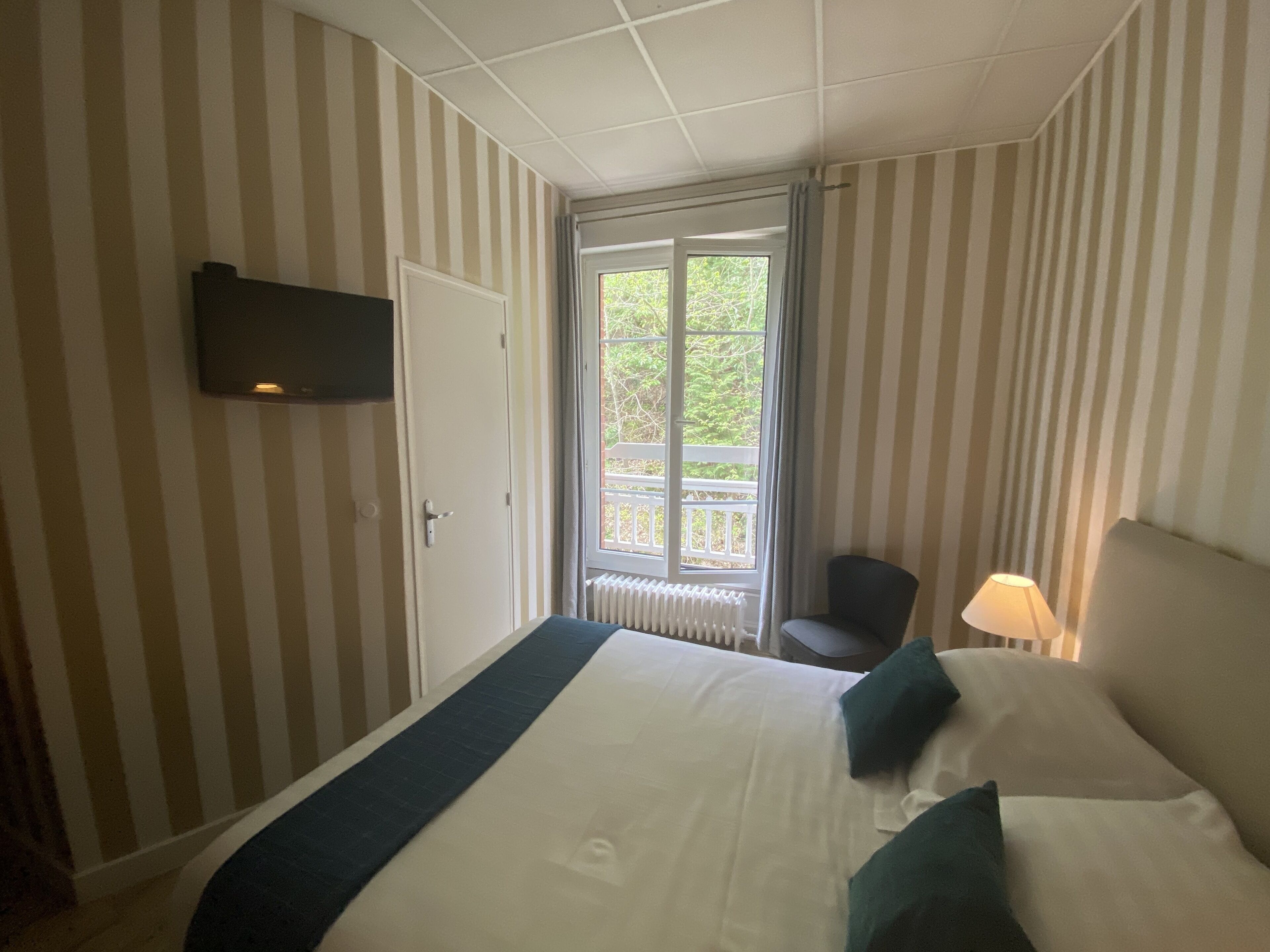 Hotelkamer met gestreepte behang, een bed met wit en donkerblauwe beddengoed, een aan de muur bevestigde televisie, een raam met uitzicht op groen en een stoel bij het raam.