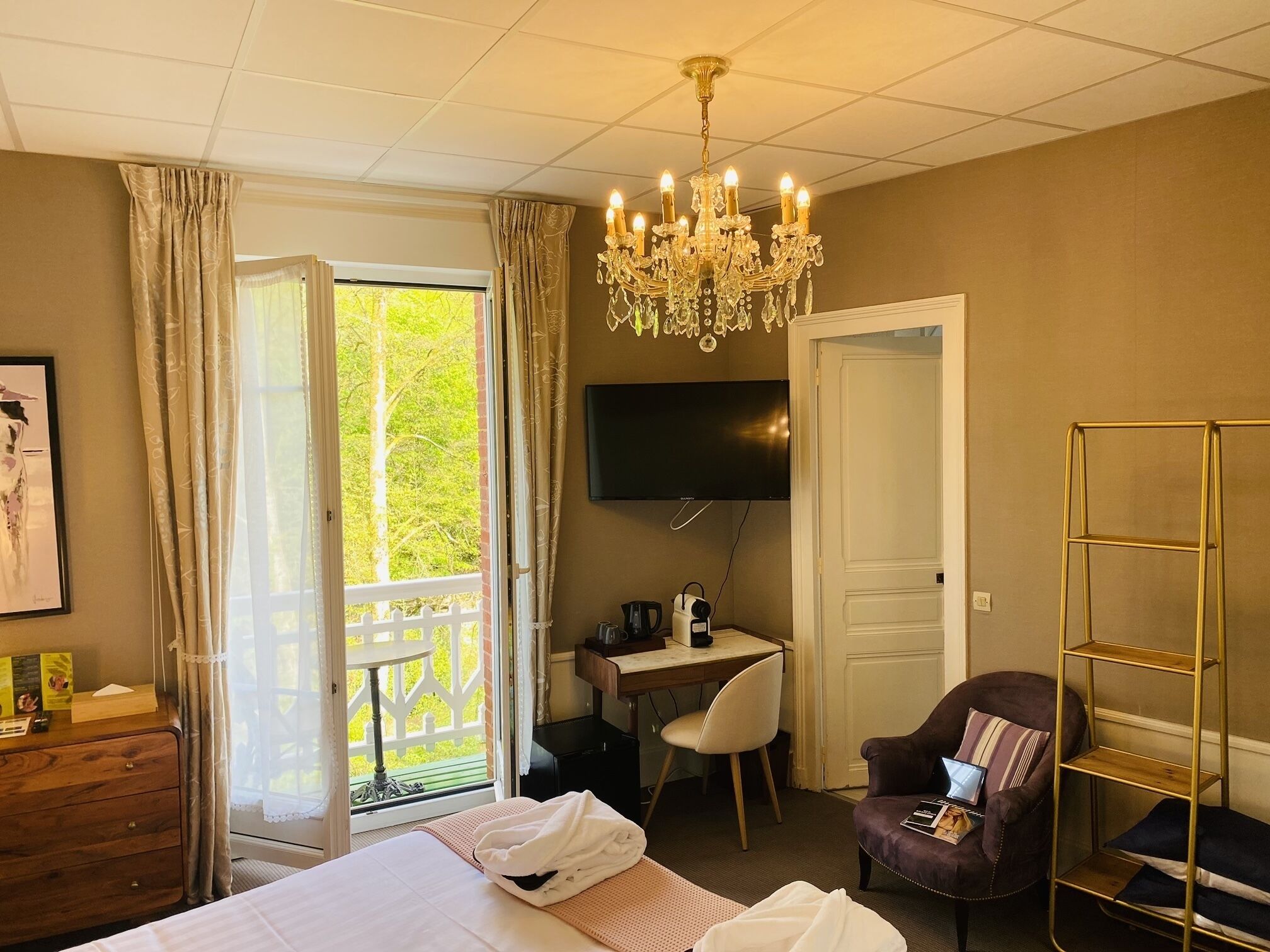 Habitación de hotel con cama, escritorio, silla, televisor y balcón con cortinas.