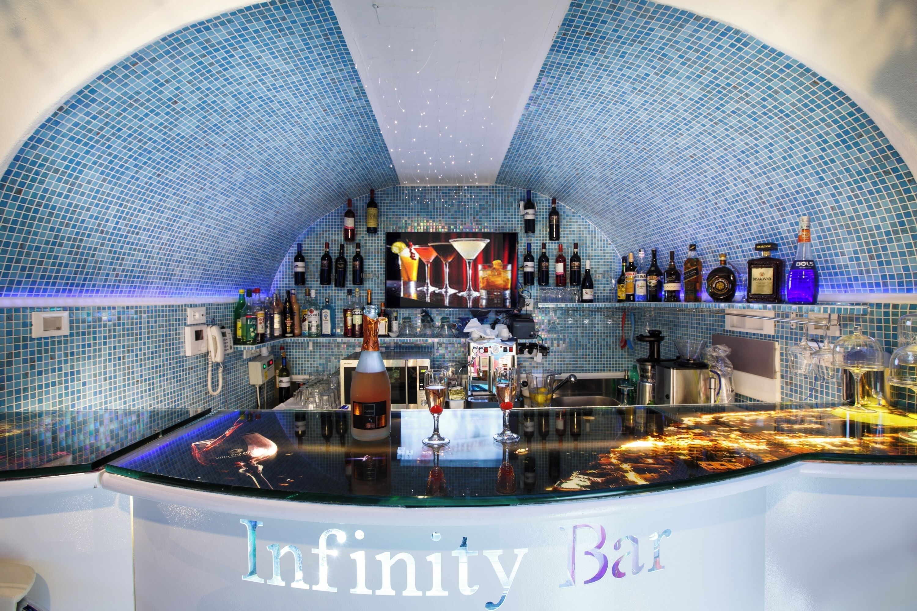 Een bar met een gebogen plafond en blauwe mozaïektegels, met een divers aanbod aan flessen en glazen.