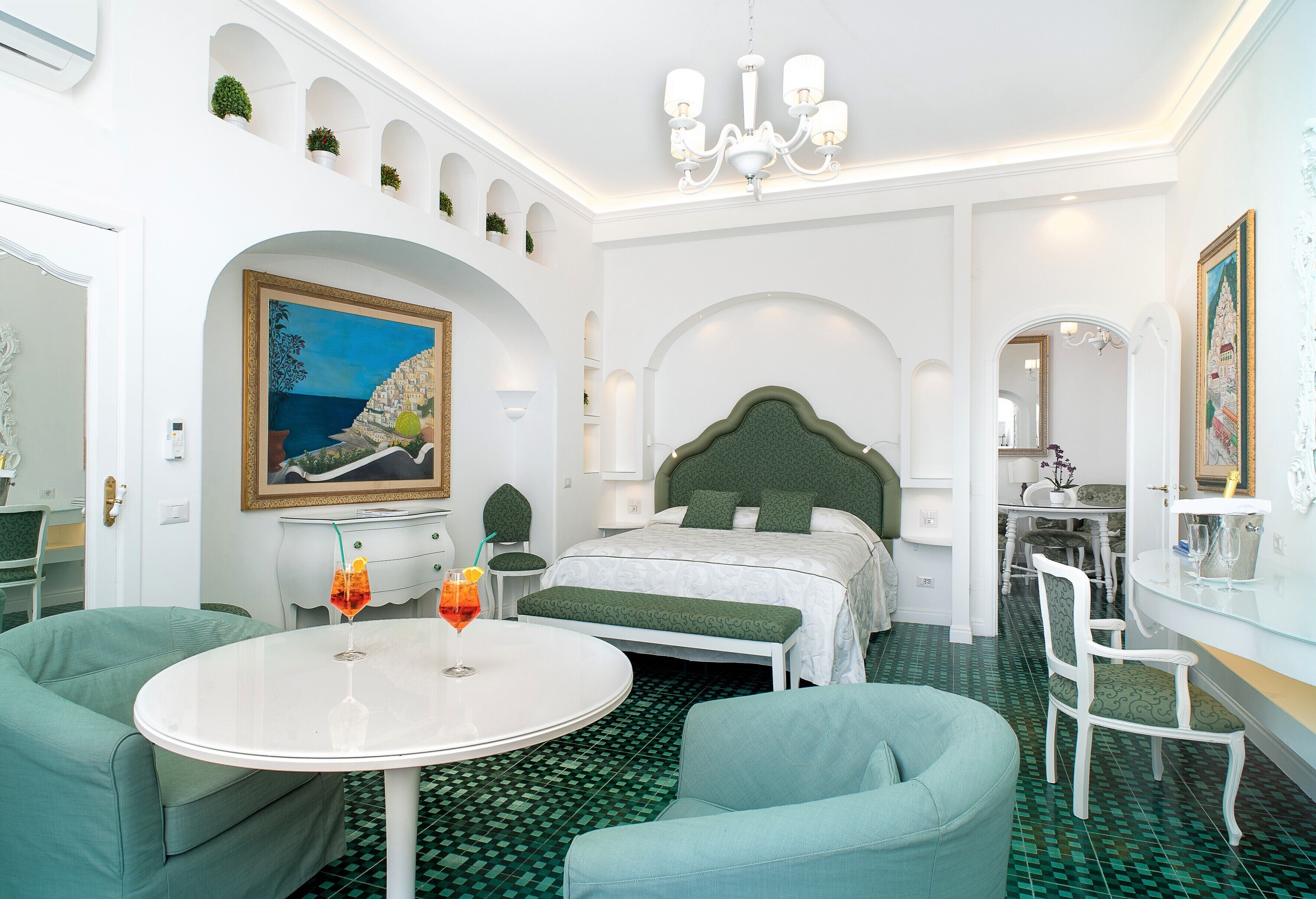 A hotel room with a bed, a round table with two drinks, a green sofa, and a dining area with chairs.