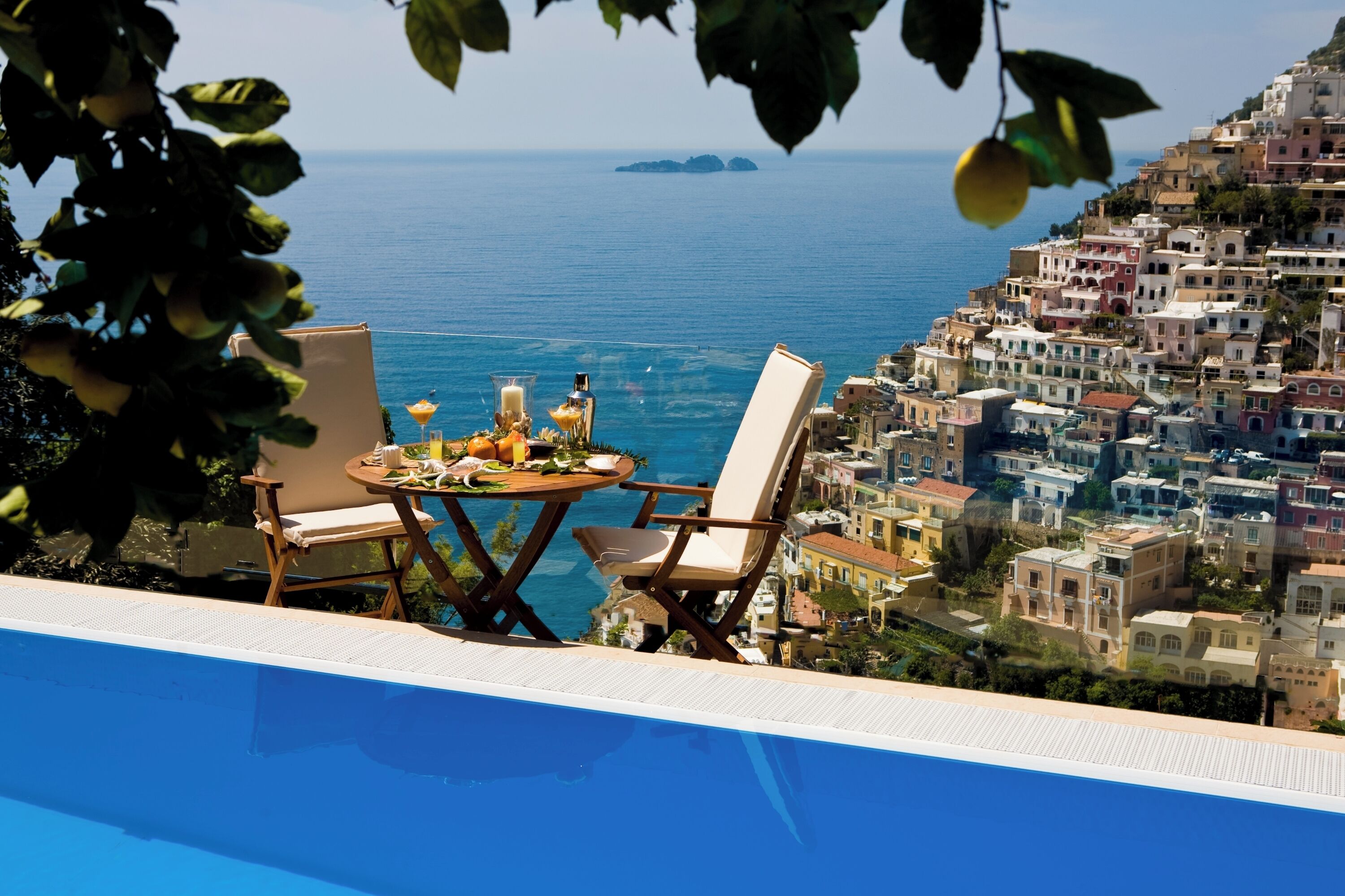 A poolside area with a table set for two, overlooking a coastal town and the sea.