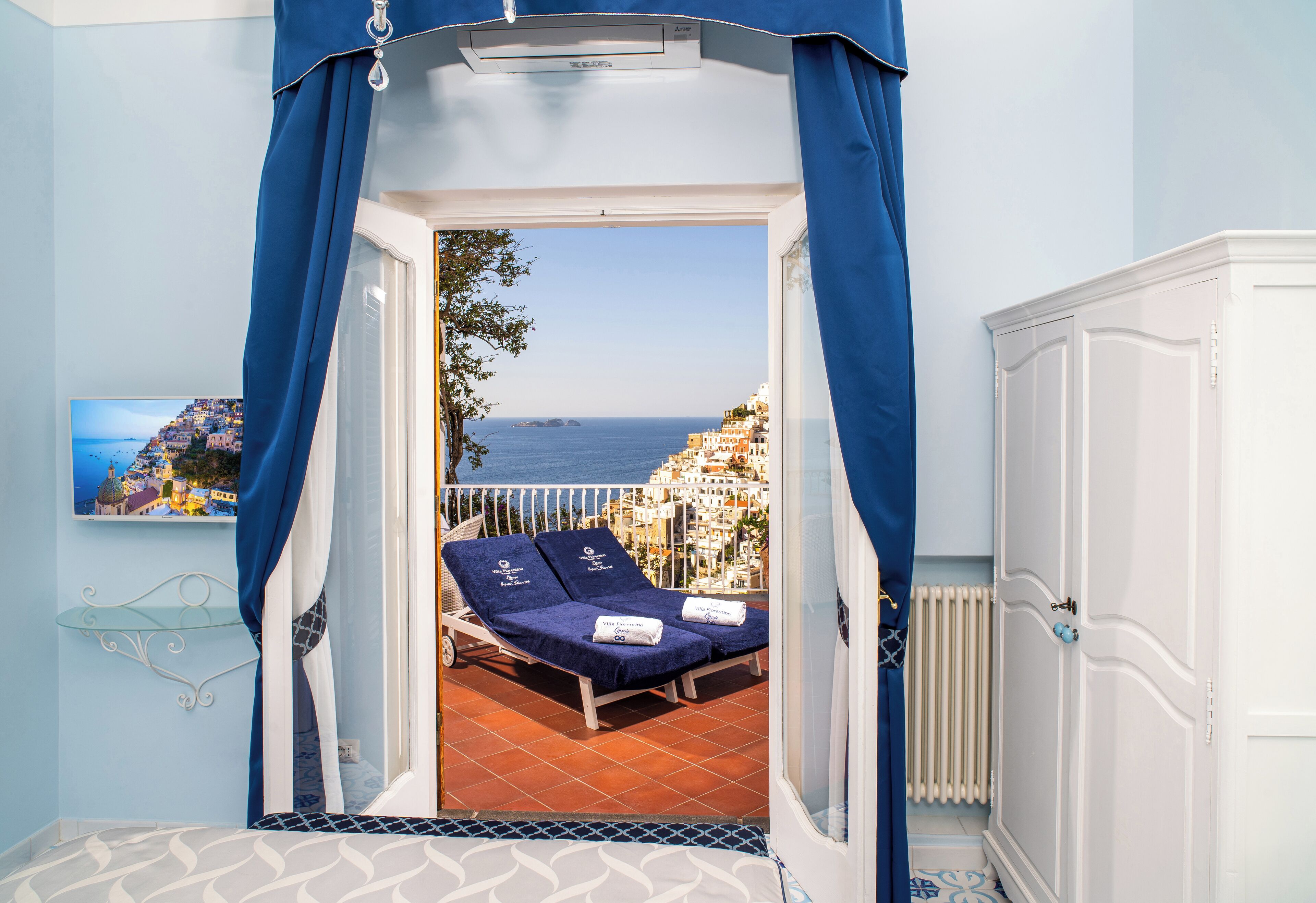Een kamer met een balkon met uitzicht op een kustplaatsje, een bed met een gestreept hoofdeinde en een witte kledingkast.