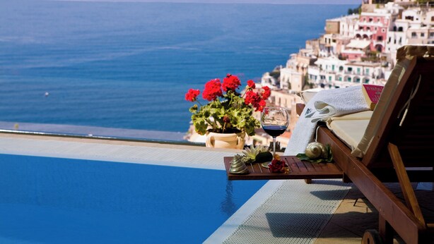 A poolside view with a chair, a glass of red wine, and a potted plant with red flowers. - VILLA FIORENTINO POSITANO  (Positano)