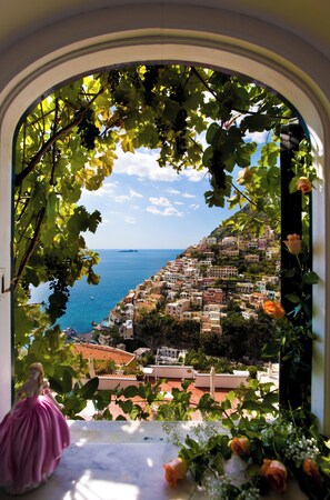 Vista a través de un arco, con un vestido rosa en primer plano, que da a un pueblo costero con edificios y el mar.