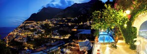 Un pueblo costero al anochecer con edificios iluminados, una zona de piscina y montañas al fondo.