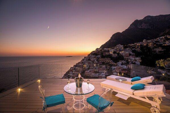 A terrace with a table, two lounge chairs, and a view of a coastal town at sunset. - VILLA FIORENTINO POSITANO  (Positano)