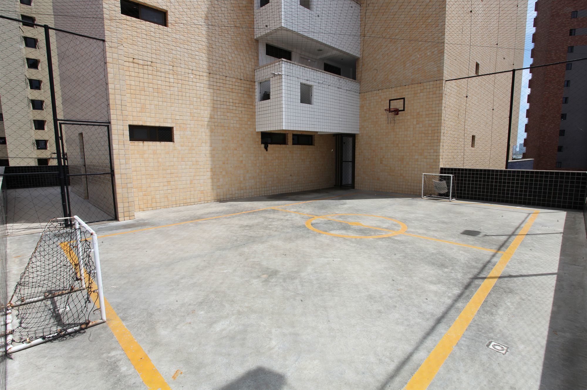 Una cancha de baloncesto al aire libre con aro, rodeada de muros de ladrillo y una entrada con portón.