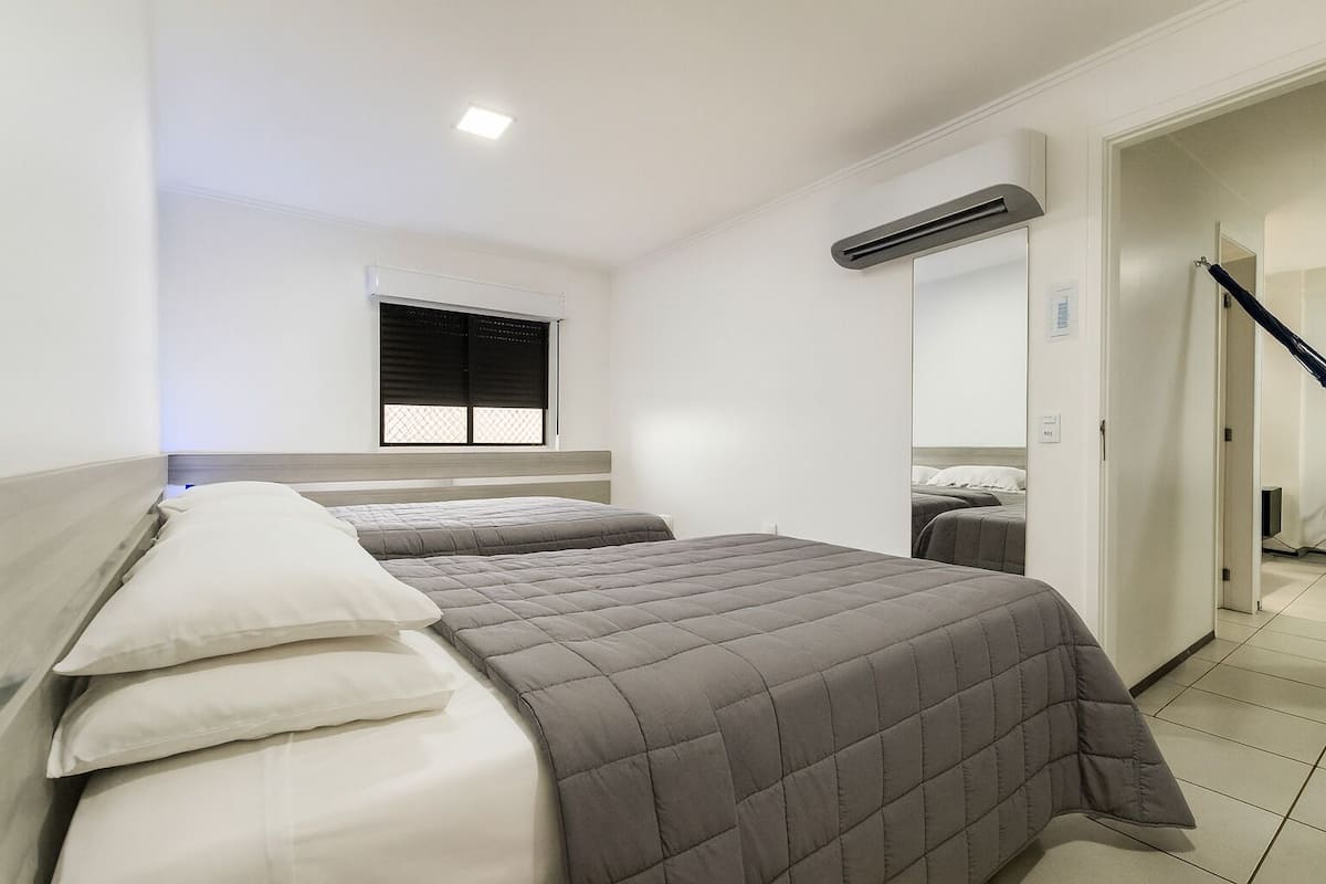 A hotel room with two beds, a window with blinds, an air conditioning unit, and a mirror on the wall.