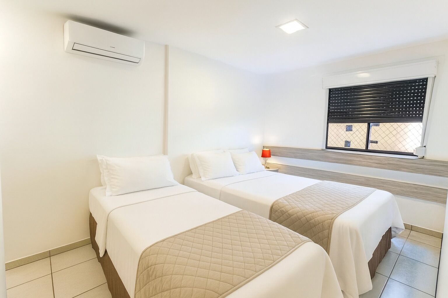 Quarto de hotel com duas camas, janela com persianas, mesa de cabeceira com abajur e aparelho de ar condicionado.
