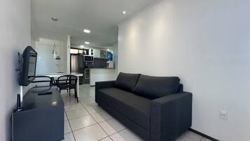 Een moderne woonkamer met een grijze bank, een flatscreen-tv aan de muur, een eettafel met stoelen en een keuken in de achtergrond.