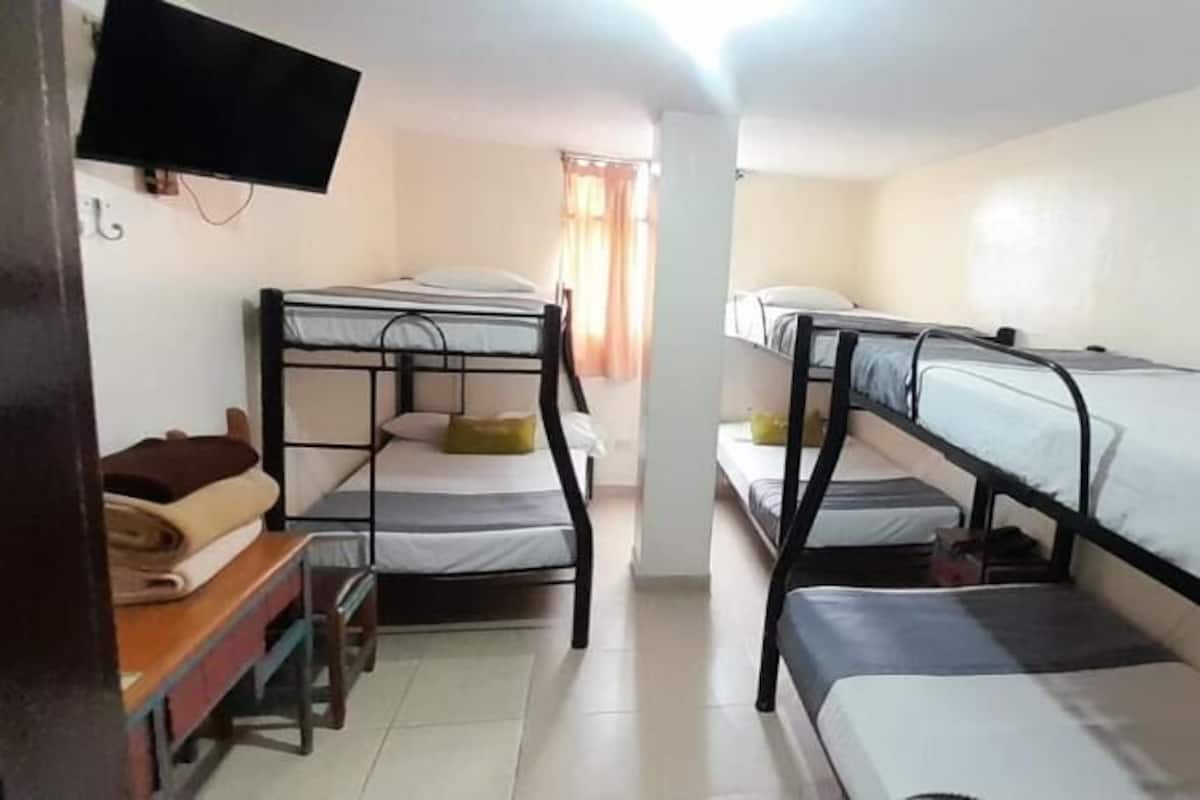 A room with bunk beds, a flat-screen TV mounted on the wall, a desk with a chair, and a window with curtains.