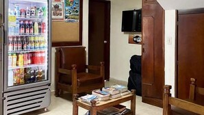 A room with a refrigerator stocked with beverages, two wooden chairs, a small wooden table with books, a TV mounted on the wall, and a door with a 'WiFi' sign.