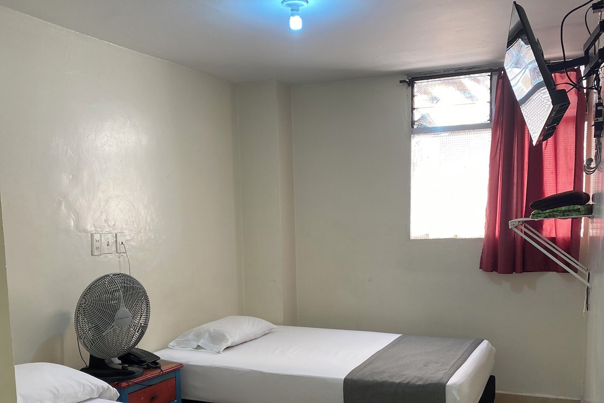 Une chambre avec deux lits, un ventilateur de plafond, un téléviseur fixé au mur et une fenêtre avec des rideaux rouges.