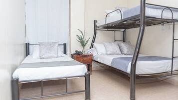 A dormitory room with two bunk beds, a bedside table with a plant, and a window with white curtains.