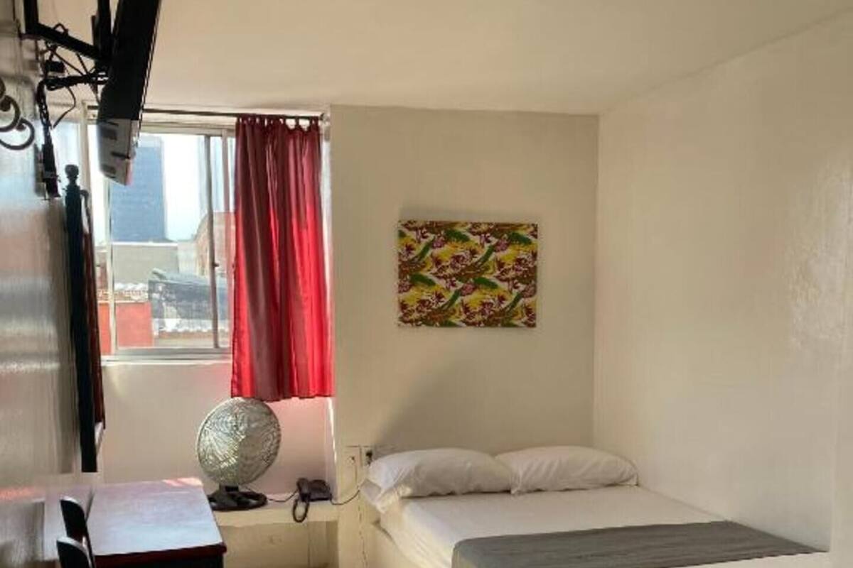 A bedroom with a bed, a desk, a chair, a window with curtains, a wall-mounted fan, and a painting.