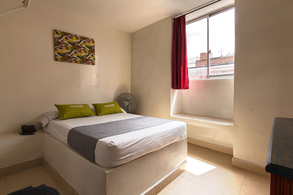 A single bed with a grey and white mattress, two pillows, a fan, and a window with a red curtain.
