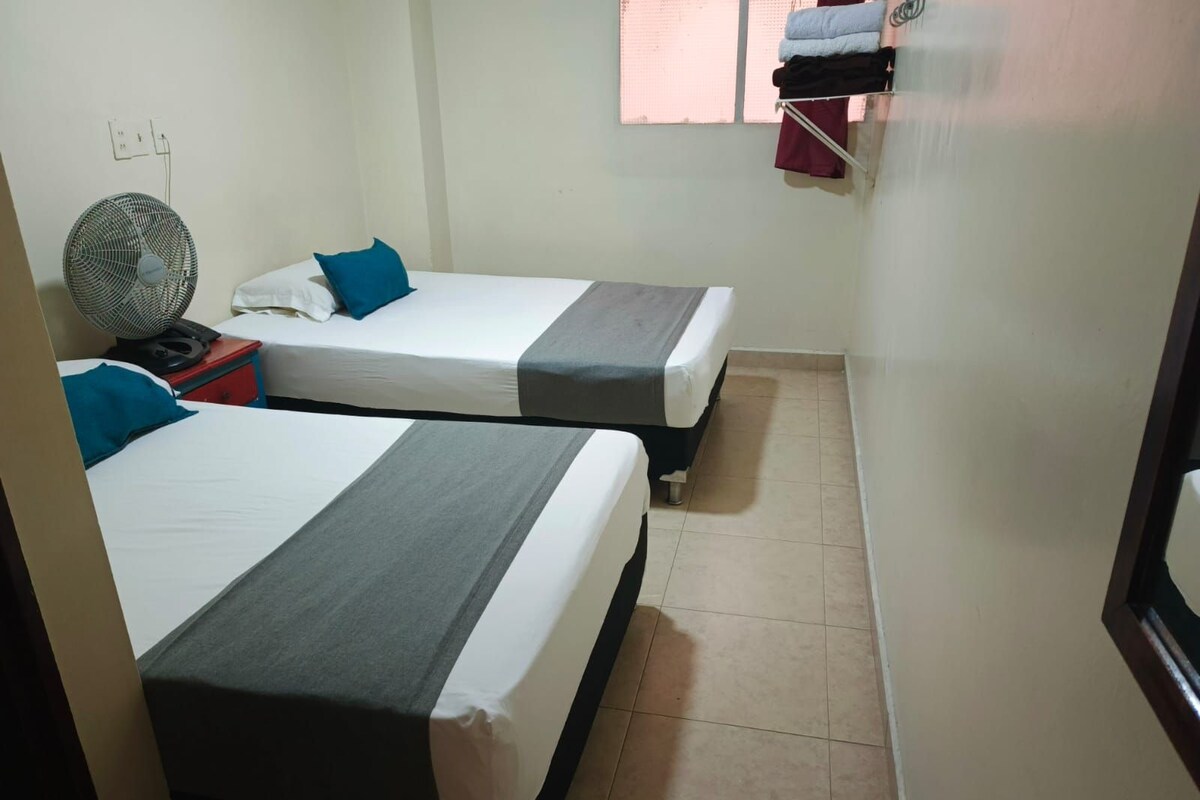A small, clean room with two single beds, a fan, and a small table.