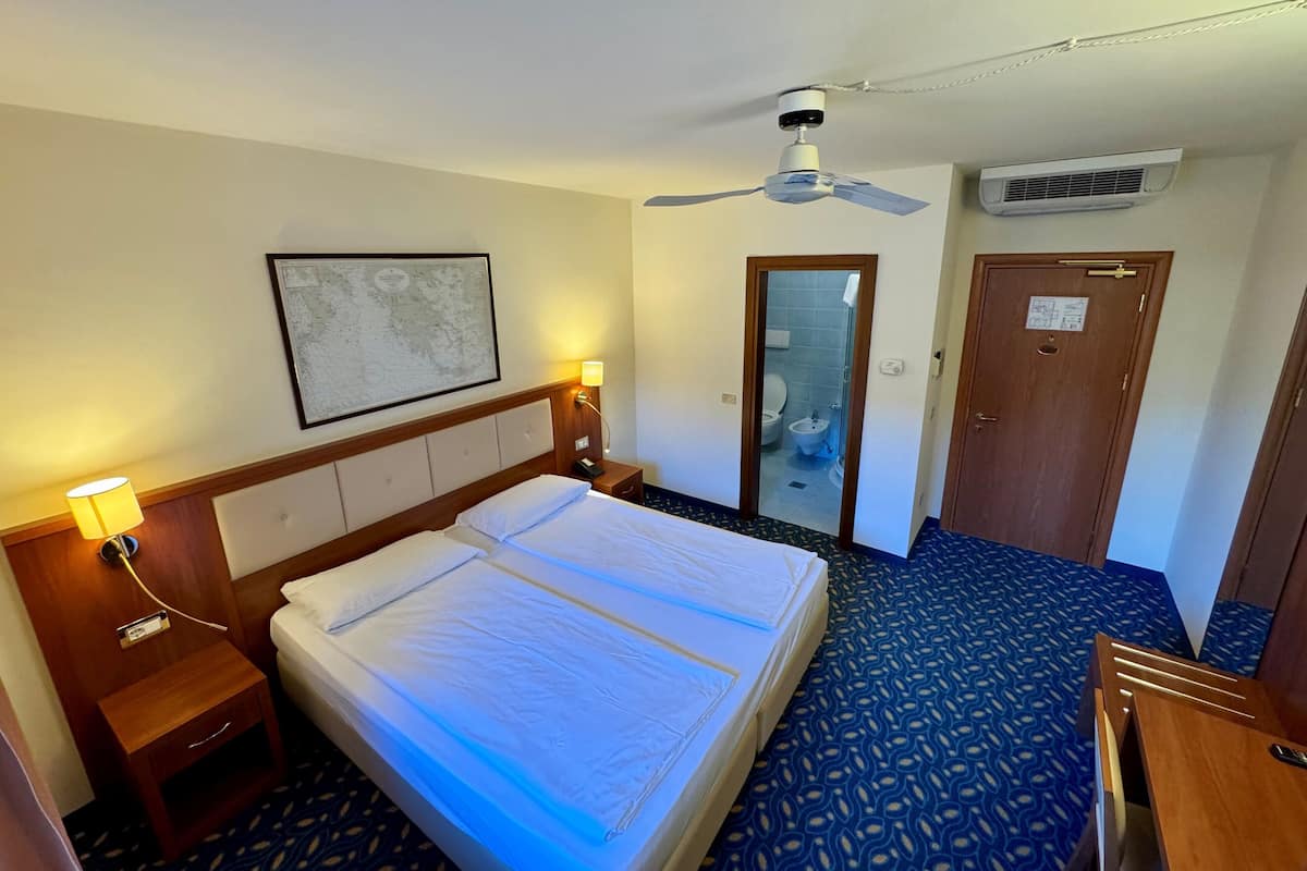 A hotel room with a bed, bedside lamps, a ceiling fan, an air conditioning unit, a bathroom door, and a framed picture on the wall.