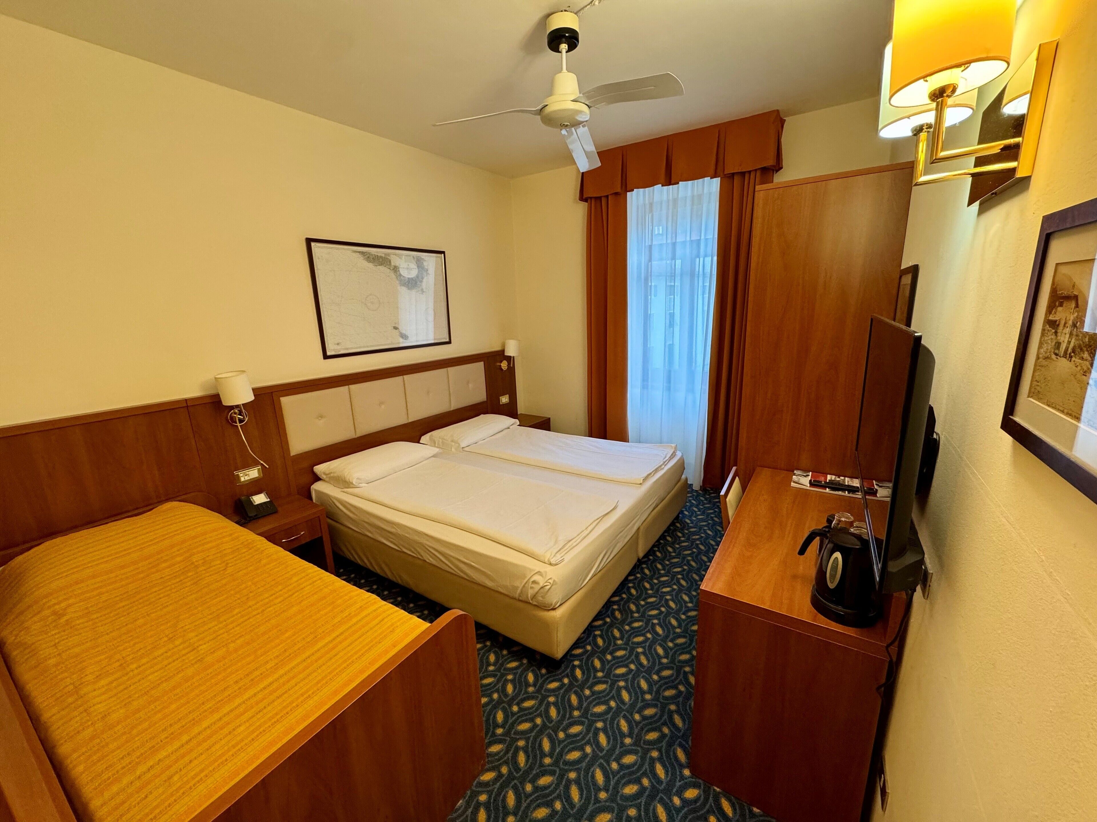 A hotel room with a double bed, a desk with a telephone, a ceiling fan, and a television.