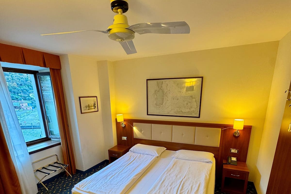 A hotel room with a bed, two bedside tables, a ceiling fan, a window with curtains, and a patterned carpet.